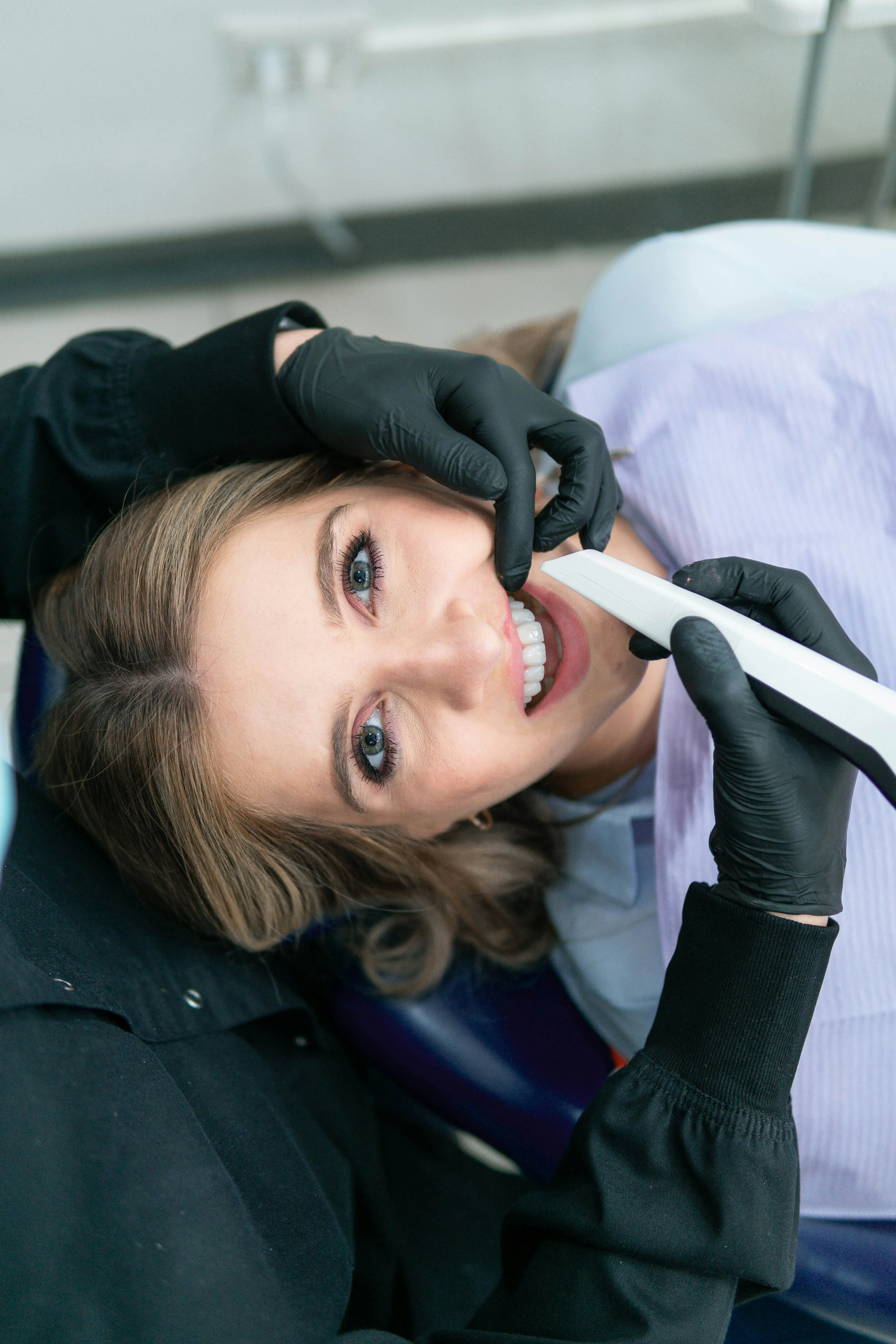 A dentist wearing black gloves uses an intraoral scanner on a smiling patient lying in a dental chair.